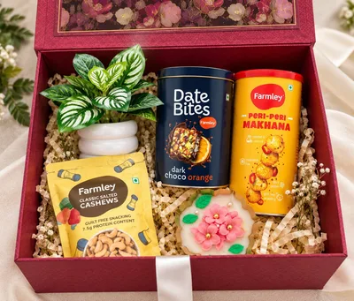 The Luxe Bloom Box open, showing an artificial plant, a floral urli candle, Farmley Date Bites, cashews and peri-peri makhana in a red gift box