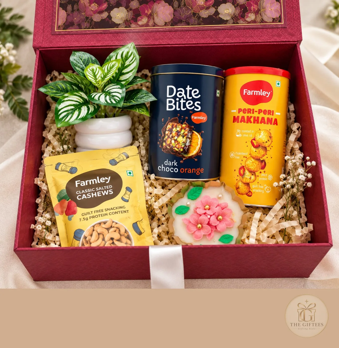 The Luxe Bloom Box, a hamper with Artificial plant in a white pot, Floral urli candle, Farmley Date Bites, dark choco orange (200g)